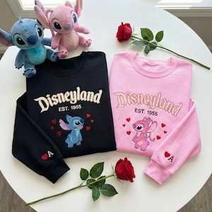 May include: Black and pink sweatshirts with embroidered "Disneyland" text and character designs. The black sweatshirt features Stitch, while the pink one showcases Angel. Both have the text "EST. 1955" and a heart with the letter "A".