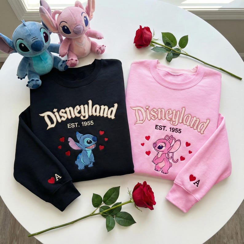 May include: Black and pink sweatshirts with embroidered "Disneyland" text and character designs. The black sweatshirt features Stitch, while the pink one showcases Angel. Both have the text "EST. 1955" and a heart with the letter "A".