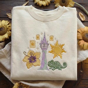 May include: Cream-colored sweatshirt with an embroidered design featuring a purple and green tower, a yellow sun, a green chameleon, and a yellow flower. The design also includes floating lanterns.