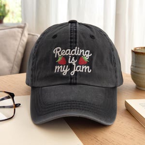 Peut inclure: Casquette de baseball noire avec l'inscription brodée "Reading is my Jam" en blanc, accompagnée de deux fraises rouges. La casquette a une visière incurvée et une sangle en tissu réglable.