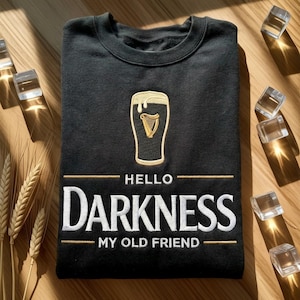 May include: Black crew neck sweatshirt featuring a gold embroidered pint of beer with a harp design. The text "HELLO DARKNESS MY OLD FRIEND" is embroidered in white below the beer glass.