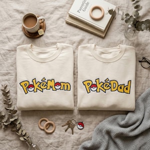 Puede incluir: Dos sudaderas color crema con los textos "PokéMom" y "PokéDad" en amarillo y rojo. Las sudaderas están sobre una superficie de tonos neutros, junto a libros, una taza de té y otros accesorios.