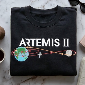 Puede incluir: Sudadera negra con la inscripción blanca "ARTEMIS II". El diseño incluye una Tierra estilizada, un satélite y la luna, conectados por una línea de trayectoria roja y naranja.