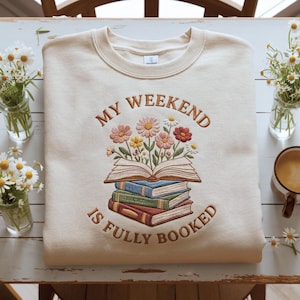 May include: Cream-coloured sweatshirt with embroidered design. The design features an open book with flowers and the text "MY WEEKEND IS FULLY BOOKED" in brown thread. The sweatshirt is folded on a white surface.