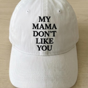 Puede incluir: Gorra de béisbol blanca con el texto bordado "MY MAMA DON'T LIKE YOU" en negro. La gorra tiene una visera curva y un botón en la parte superior. El sombrero está hecho de una tela suave y texturizada.