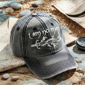 May include: A distressed gray baseball cap with the text "I am no man" in white script, along with a sword and leaf design. The cap has a curved brim and a classic six-panel design.
