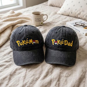 Puede incluir: Dos gorras de béisbol negras con las palabras "PokéMom" y "PokéDad" bordadas en amarillo. Las gorras están sobre una cama con una manta de color neutro. Una taza con "Cozy Home" está en el fondo.