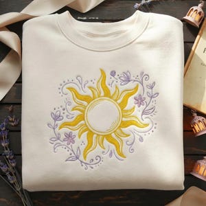 May include: Cream-colored sweatshirt featuring a yellow sun design with intricate details. The sun is surrounded by a wreath of purple flowers and vines. The design is embroidered on the front of the sweatshirt.