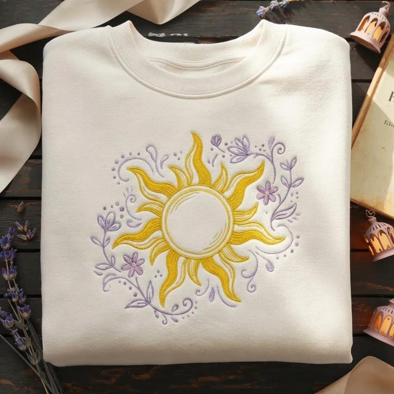 May include: Cream-colored sweatshirt featuring a yellow sun design with intricate details. The sun is surrounded by a wreath of purple flowers and vines. The design is embroidered on the front of the sweatshirt.