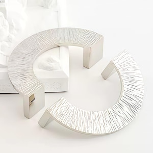 Puede incluir: Un modelo arquitectónico blanco y una escultura floral blanca se exhiben con dos tiradores de armario plateados. Los tiradores tienen un diseño lineal texturizado. El modelo arquitectónico representa un edificio con múltiples cúpulas y una escalera.