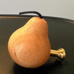 Può includere: Un pomello per mobili a forma di pera realizzato in legno chiaro con venature visibili. Il pomello presenta un piccolo stelo nero ricurvo sulla parte superiore ed è fissato a una base metallica cilindrica color ottone per il montaggio.
