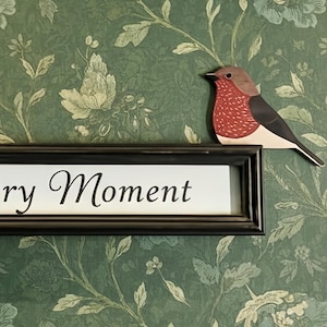 May include: A framed sign with the words "Cherish Every Moment" in elegant script. The sign is black and mounted on a green floral wallpaper. A wooden bird decoration is perched above the frame.