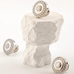 Op de afbeelding: Zilveren kastknoppen met een decoratief cirkelvormig ontwerp. De knoppen hebben een centraal bloemmotief omgeven door geometrische vormen. Drie knoppen worden getoond op een wit, getextureerd oppervlak, waarvan één knop op een gestapelde steenachtige structuur rust.