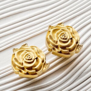 May include: Two gold-colored metal rose-shaped decorative knobs or pulls. Each piece features a detailed floral design with layered petals and a leaf accent. The dimensions are clearly printed as 0.98 by 1.38 inches with a weight of 67 grams.