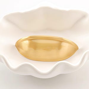 Peut inclure: Une poignée de meuble incurvée de couleur or repose à l'intérieur d'un plat en céramique blanc à bord festonné. La poignée a une surface lisse et réfléchissante. Le plat a une forme ondulée et organique, créant un contraste avec la forme de la poignée.