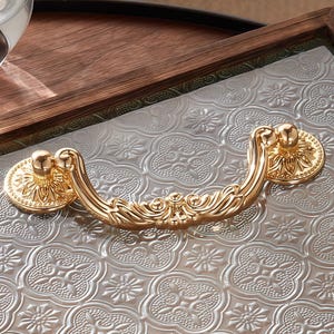 Könnte beinhalten: Nahaufnahme eines dekorativen Tabletts mit einem goldfarbenen Griff. Der Griff weist ein verziertes Design mit floralen Details auf. Das Tablett hat eine transparente, gemusterte Oberfläche und einen Holzrahmen.