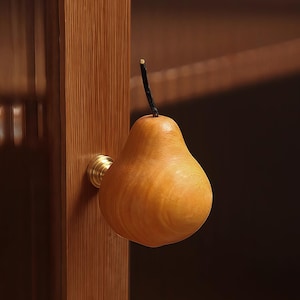 May include: A wooden pear-shaped cabinet knob with a brass base and a dark stem. The knob is attached to a wooden cabinet door. The pear is a light brown color.