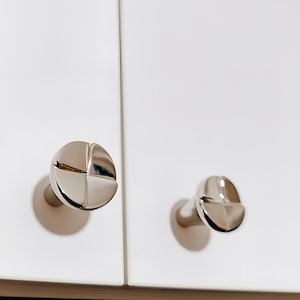 May include: Two polished chrome cabinet knobs with a screw head design. The knobs are mounted on a white cabinet door. The knobs have a circular base and a rounded, grooved head.