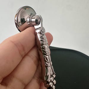Op de afbeelding: Een meubelgreep van gepolijst chroom met een ronde montagebasis en een scharnierende, langwerpige handgreep. De handgreep heeft een gestructureerd, bladachtig patroon over de lengte en eindigt in een cluster van kleine, ronde bolletjes.