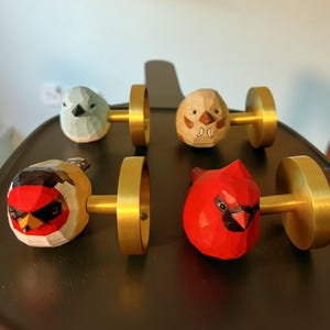 May include: Four wooden cabinet knobs, each shaped like a bird. The birds are painted in shades of red, blue, and beige. They are mounted on gold-coloured metal bases, suitable for drawers or cupboards.