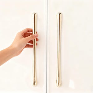 Może przedstawiać: Para złotych uchwytów do szafek o eleganckim, wydłużonym kształcie. Jeden uchwyt jest trzymany przez dłoń, prezentując gładkie, wypolerowane wykończenie. Uchwyty są zamontowane na białych drzwiach szafki, oferując nowoczesną estetykę.