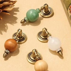 Op de afbeelding: Een verzameling decoratieve kastknoppen met messing hardware. De knoppen hebben verschillende ontwerpen, waaronder een groene jade bol, een lichtbruine houten bol, een witte bol en een lichtbruine houten bol met een decoratieve band.