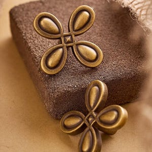 Op de afbeelding: Twee antieke messingkleurige kastknoppen met een vierbladontwerp. De knoppen zijn geplaatst op een getextureerd, bruin oppervlak. De knoppen hebben een vierkant midden en een vintage esthetiek.