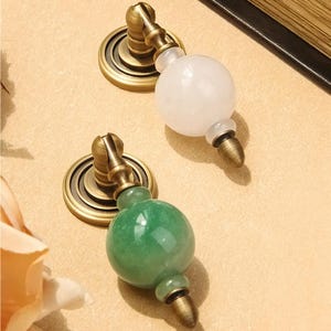 May include: Two cabinet knobs with a vintage aesthetic. One knob features a white, spherical stone, while the other has a green stone. Both have antique brass-colored hardware and circular backplates, offering a classic look for furniture.