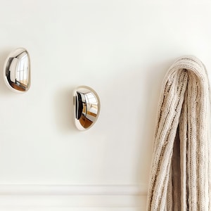 May include: Two polished chrome wall hooks with a teardrop shape are mounted on a white wall. A beige knit scarf hangs from one of the hooks. The hooks reflect the surrounding environment.