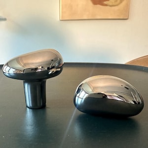 Könnte beinhalten: Zwei glänzende, dunkelgraue Dekorationsgegenstände. Eines ist pilzförmig mit abgerundeter Oberseite und zylindrischem Sockel. Das andere ist ein glatter, oval geformter Stein. Beide haben eine reflektierende, metallische Oberfläche.