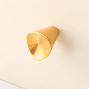 Puede incluir: Un pomo de mueble de color dorado con un acabado metálico cepillado y liso. El pomo presenta una forma asimétrica distintiva, similar a un triángulo con bordes redondeados, que se estrecha hacia la base donde se fija a la superficie plana color crema.