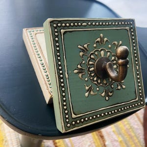 Puede incluir: Dos perchas decorativas de pared. Una es de color verde azulado con un gancho de bronce y un diseño floral ornamentado. La otra es de color blanquecino con un borde de cuentas. Ambas son cuadradas y de estética vintage.