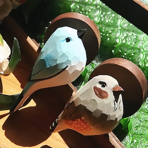 Peut inclure: Deux sculptures d'oiseaux en bois. L'un est bleu clair avec un bec noir, l'autre est blanc et marron avec une poitrine tachetée rouge-orangé. Les oiseaux sont perchés sur une branche en bois, avec un arrière-plan vert flou.