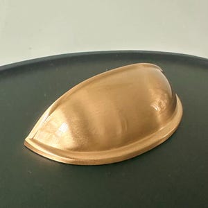 May include: A gold-coloured, cup-style cabinet pull with a curved design. The hardware has a smooth, reflective surface and is likely made of metal. The pull is designed to be mounted on a drawer or cabinet door for easy opening.