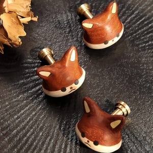 Könnte beinhalten: Drei holzgeschnitzte Schrankknöpfe in Fuchskopfform mit Messingstiften. Die Knöpfe sind braun mit weißen Akzenten an Ohren und Gesicht. Die Knöpfe sind auf einer dunklen, strukturierten Oberfläche angeordnet.
