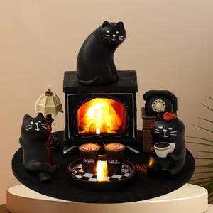 May include: A whimsical black cat-themed miniature scene. It features a fireplace with a glowing fire, a lamp, a telephone, and several black cat figurines. Two coffee cups sit on a checkered tray. The scene is set on a black felt mat.