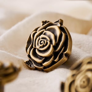 May include: A bronze-coloured, rose-shaped decorative knob. The knob features detailed petals and a central spiral, set against a soft, off-white fabric background. The hardware is likely for furniture or cabinetry.