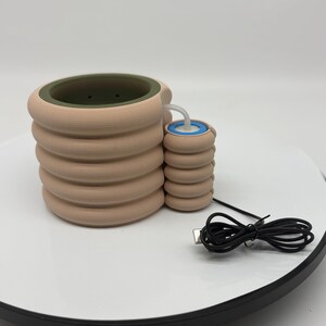 Puede incluir: Un objeto cilíndrico beige apilado con un cilindro más pequeño y conectado. El cilindro más grande tiene un interior verde. Un componente azul y blanco es visible en el cilindro más pequeño. Un cable USB negro está conectado.