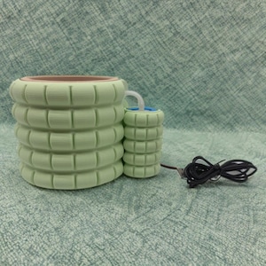 Puede incluir: Un difusor de aromaterapia cilíndrico verde claro con un componente más pequeño a juego. El difusor tiene un diseño texturizado y apilado. Un cable de alimentación negro está conectado al componente más pequeño. La parte superior del difusor es marrón.