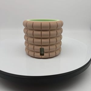 Puede incluir: Una maceta de cerámica de color beige, de forma cilíndrica, con un diseño de ladrillos apilados en relieve. El borde interior es verde claro. La maceta está sobre una superficie blanca con un borde negro. Tiene una pequeña abertura rectangular.