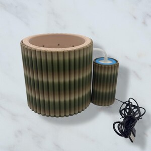 Puede incluir: Un juego de macetas cilíndricas de dos piezas, con estampado de camuflaje. La maceta más grande tiene un interior beige y un exterior acanalado en tonos verdes y marrones. Un cilindro más pequeño a juego con el interior azul está conectado por un tubo blanco.