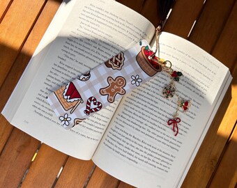 May include: A festive bookmark with a gingerbread man, candy cane, and waffle design, resting on an open book. The bookmark has a gold chain with Christmas-themed charms, including a snowflake and a red bow. The book is open on a wooden surface.