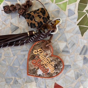 May include: A keychain accessory featuring a heart-shaped patch with the embroidered text Pumpkin Spice in orange and yellow thread. The design includes a leopard print pouch, a dark brown tassel, a braided cord, and small fuzzy brown beads.