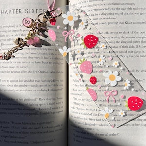May include: A clear acrylic bookmark with strawberry, daisy, and bow designs. It has a pink tassel and a charm with a pink letter "S" and pink beads. The bookmark is placed in an open book.
