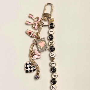 Pode incluir: Chaveiro dourado com vários pingentes, incluindo um laço rosa, uma xícara de café, um livro com um coração, uma borboleta e um coração xadrez em preto e branco. O chaveiro soletra "IANNALOPEZ" com contas de letras pretas e brancas.