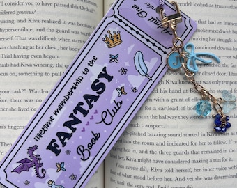 May include: A lavender bookmark with the text "FANTASY Book Club" and decorative elements like a dragon, crown, and feather. It has a tassel and a charm with a key and the letter "J".