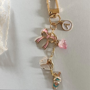 May include: Gold-tone keychain with a lobster clasp, featuring a pink bow, a butterfly, a flower, and a drink charm. The keychain also includes a charm with the letter "A".