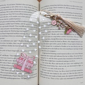 Op de afbeelding: Een bladwijzer met een heldere, rechthoekige bedel, een goudkleurige sluiting, een kwast en roze bedels. De bladwijzer ligt op een open boek met tekst en een roze en witte speld met de tekst "She Mirrya" en een stapel boeken.