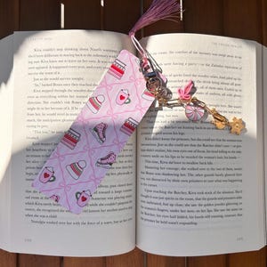 May include: A pink bookmark with a repeating pattern of winter hats, ice skates, books, and mugs of hot chocolate. The bookmark is attached to a book with a tassel and decorative charms.