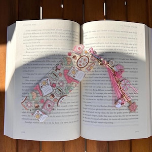 Könnte beinhalten: Ein dekorativer Lesezeichen mit einem rosa Quasten und Anhänger, das in einem offenen Buch liegt. Das Lesezeichen hat ein skurriles Design mit Illustrationen von Büchern, Teetassen und Gebäck in Rosa- und Beigetönen. Das Buch ist auf einer Holzoberfläche geöffnet.